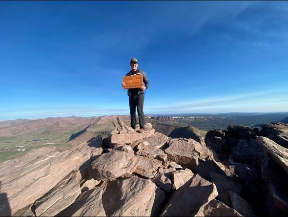 Justin Manco on the summit of Kings Peak, the highest point in Utah, holding the summit marker against a blue sky and desert horizon.